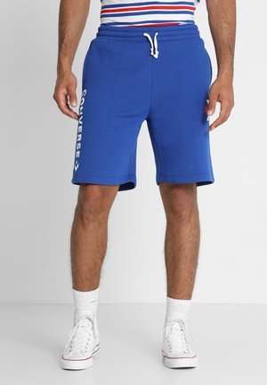 Homme portant un short Converse bleu avec logo sur le côté, des baskets montantes blanches et des chaussettes blanches, debout devant un fond uni.