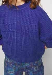 Lila stickad tröja med en texturerad yta och rund hals. Matchas med mönstrade blå och gröna byxor med grafiska mönster.