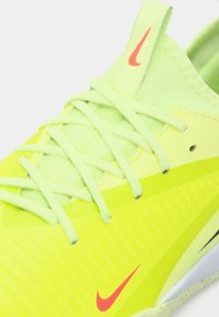 Jasnożółty but sportowy z gładką powierzchnią, teksturowanymi akcentami i jasnozielonymi sznurowadłami. Zawiera małe czerwone logo Nike.