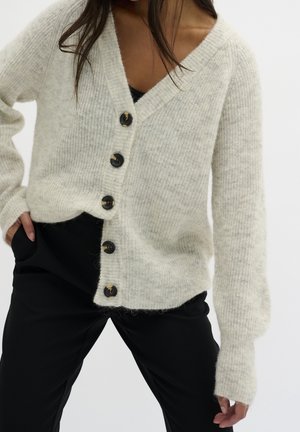 Femme portant un cardigan boutonné doux, gris clair, avec un col en V profond, sur un pantalon noir, mains dans les poches sur un fond blanc uni.