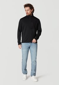 Schwarze Zip-Jacke mit hohem Kragen, aus weichem Material gefertigt, mit einer Brusttasche. Kombiniert mit hellblauen Jeans und weißen Turnschuhen.