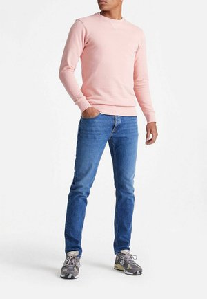 Man draagt een lichtroze shirt met lange mouwen, blauwe spijkerbroek en grijze sneakers, staat met één hand in zijn zak tegen een witte achtergrond.