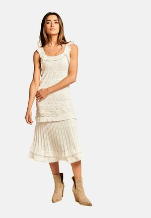Weißes Strickkleid mit quadratischer Ausschnitt, Rüscheträgern, strukturierten Mustern und geschichteten Falten am Saum, kombiniert mit braunen Knöchelstiefeln.