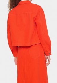 Žena s kudrnatými vlasy, oblečená v jasně oranžové zkrácené bundě a odpovídající sukni s vysokým pasem, zobrazená zezadu.