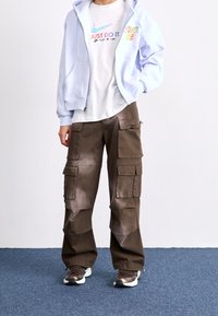 Ljusblå zip-up huvtröja, vit t-shirt med flerfärgad "JUST DO IT" tryck, bruna cargobyxor med fickor och mörka sneakers.