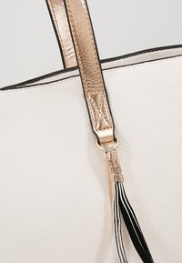 Bolso de mano de cuero blanco texturizado con una correa metálica color oro rosado, que cuenta con un contorno negro y acentos decorativos de flecos a rayas.