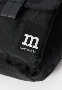 Sac à dos en tissu noir avec un motif texturé, un rabat avant avec boucle et une étiquette Marimekko avec un logo blanc distinct.