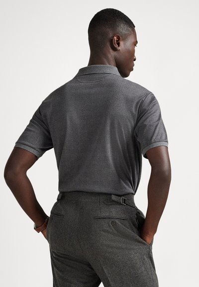Άνδρας που φοράει γκρι καρό κοντομάνικο πόλο πουκάμισο, μπαλαρισμένο μέσα σε σκούρο γκρι παντελόνι ραμμένο, στέκεται με τα χέρια στις τσέπες, με την πλάτη στραμμένη.