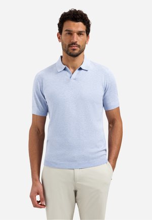Man draagt een lichtblauw poloshirt met korte mouwen en beige broek, staat met één hand in de zak tegen een effen achtergrond.