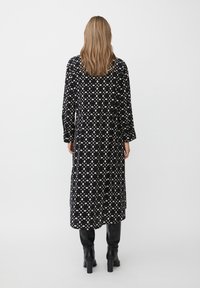 Langes schwarzes Kleid mit cremefarbenen Tupfen, das lange Ärmel, eine taillierte Taille und eine gerade Silhouette aufweist. Getragen mit schwarzen Absatzstiefeln.