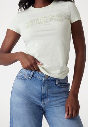 Femme portant un t-shirt Guess vert clair avec logo brodé et un jean bleu taille haute, tenant l'ourlet du t-shirt d'une main.