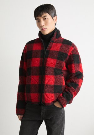 Veste en polaire avec un motif carreaux rouges et noirs, col montant, fermeture éclair à l'avant et deux poches latérales, texturée, coupe décontractée.