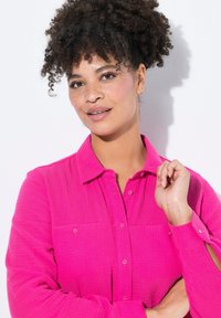 Fuchsia Hemd mit strukturiertem Stoff, langen Ärmeln und zwei Fronttaschen. Modell posiert mit einer erhobenen Hand vor einem weißen Hintergrund.