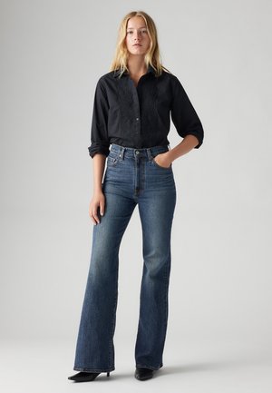 Black button-up shirt with lace detailing, paired with high-waisted blue flared jeans, and black heeled shoes. Smooth fabric and fitted design.