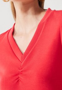 Top rojo de cuello en V con un detalle fruncido en el centro. Hecho de una tela suave, presenta una forma ajustada que realza el escote.