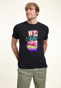Camiseta negra de manga corta con texto colorido en estilo graffiti que incluye los diseños de "NYC", "LA", "París" y "Londres". Material suave y ajustado.