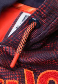 Gestructureerde stof in donkerblauw met een rood en oranje ruitpatroon; heeft een decoratieve oranje trekkoord en contrasterende navy hardware.