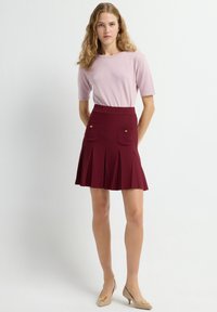 Bordeaux geplooide rok met twee voorzakken, gouden knopen en een aansluitende tailleband. Gecombineerd met een lichtroze top met korte mouwen en beige hakken.
