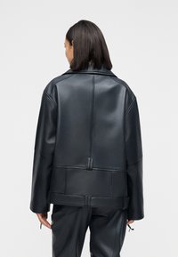 Γυναίκα με μαλλιά μέχρι τους ώμους φοράει μια μαύρη υπερμεγέθη δερμάτινη ζακέτα και αντίστοιχο δερμάτινο παντελόνι, εμφανιζόμενη από πίσω σε απλό φόντο.