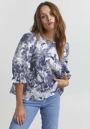 Weiße Bluse mit blauem Blumenmuster, rundem Ausschnitt und Rüschenschultern. Kombiniert mit hellblauen Jeans, die einen lässigen Sitz zeigen.