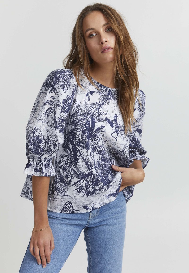 Blusa bianca con motivo floreale blu, scollatura rotonda e maniche a volant. Abbinata a jeans azzurri chiari, che mostrano una vestibilità rilassata.