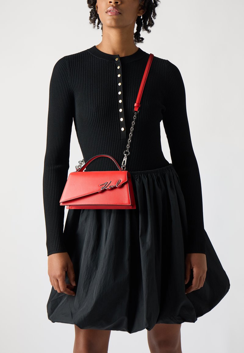 Borsa strutturata rossa con manico superiore e tracolla a catena, con un accento del logo. Il modello indossa un vestito lungo a maniche lunghe a coste nero con gonna svasata.