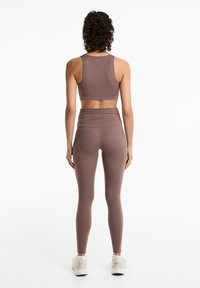 Sutien sport și colanți până la talie dintr-un material maro desaturat. Are un design elegant, cu o textură netedă și detalii plisate la talie.