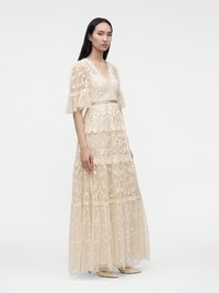 Femme debout portant une longue robe en dentelle beige avec des motifs floraux et des manches courtes transparentes à volants sur un fond blanc uni.