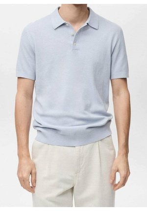 Homme portant un polo bleu clair à manches courtes rentré dans un pantalon beige, debout avec les bras détendus le long du corps.