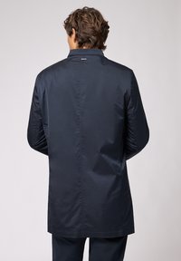 Mørkeblå, langline letvægts vandafvisende jakke med stående krave, glat tekstur og diskret logo detaljer på bagsiden.