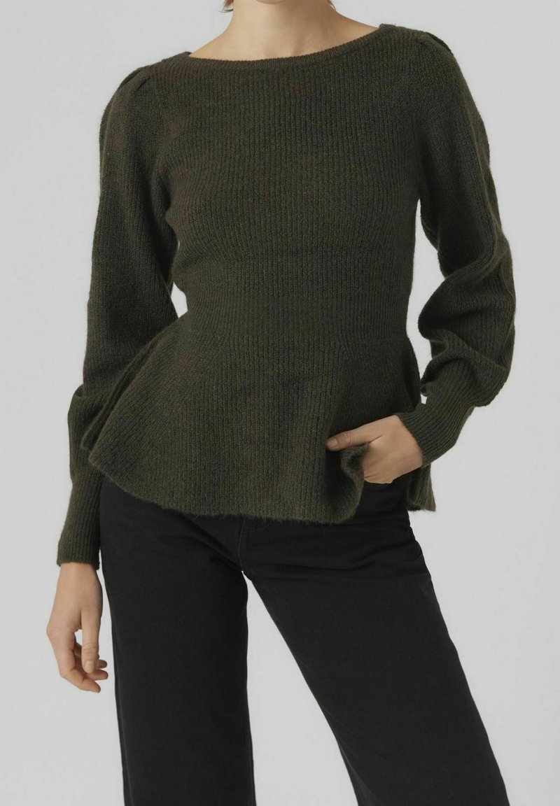 Donna in un maglione a costine verde scuro con maniche a sbuffo e pantaloni neri, con una mano in tasca, contro uno sfondo grigio semplice.