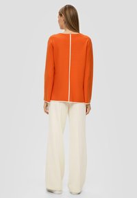 Oranje langarmig shirt met een middennaad en witte accenten, gecombineerd met crèmekleurige, brede ribbroek gemaakt van een zachte stof.