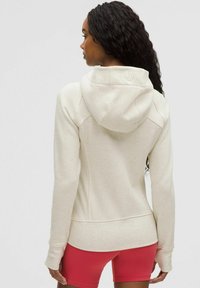 Krem dukserica s kapuljačom, teksturirana tkanina, prilagođen dizajn i otvori za palčeve. Nosena s crvenim šortsevima, ističući elegantnu siluetu.