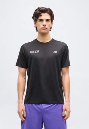 Νέος άνδρας με σκουρόχρωμα μαλλιά, φορώντας μαύρο T-shirt New Balance με λογότυπα και μοβ σορτς, στέκεται μπροστά σε μονόχρωμο ανοιχτό φόντο.