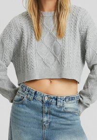 Vrouw in een lichtgrijze kabelgebreide cropped sweater en hoge taille blauwe spijkerbroek, met een blote buik en handen op de heupen.