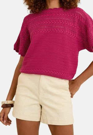 Kvinde iført en fuchsia strikket kortærmet sweater med hulmønstre og beige højtaljede shorts mod en ensfarvet baggrund.