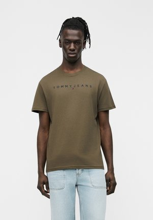 Sötét bőrű, dreadlock-frizurás férfi, olívazöld Tommy Jeans pólóban és világoskék farmerban, egyszerű háttér előtt áll.