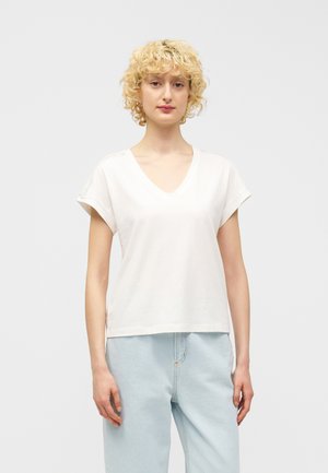 Vrouw met kort krullend blond haar, gekleed in een wit T-shirt met V-hals en korte mouwen en lichtblauwe jeans, staand tegen een effen achtergrond.