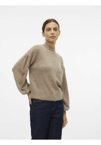 Ljusbrun stickad tröja med hög hals och lösa, puffärmar i långt format. Bärs med mörka denimjeans. Mjuk textur och avslappnad design.