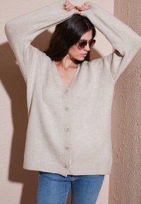 Cardigan lavorato a maglia beige con scollatura a V, maniche lunghe e chiusura a sei bottoni. Orlo e polsini a coste; texture morbida e vestibilità rilassata.