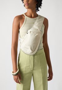 Beige sling bag featuring a smooth, padded design with a zip pocket and logo detail. Worn on the shoulder, paired with green trousers.