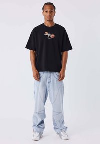 Camiseta negra oversized con diseño gráfico, combinada con jeans baggy de color azul claro que presentan patrones de costura curvada y deportivas blancas.