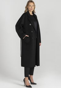 Czarny wełniany długi płaszcz o podwójnym zapięciu, z dużymi klapami, bocznymi kieszeniami i luźnym krojem, w zestawieniu z czarnymi spodniami i szpilkami.