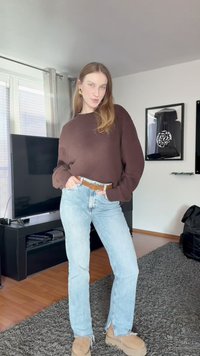 Νεαρή γυναίκα με μακριά μαλλιά, φορώντας καφέ πουλόβερ, ανοιχτό μπλε τζιν και μπεζ παπούτσια, στέκεται σε ένα μοντέρνο σαλόνι.