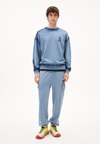 Man in een lichtblauwe sweatshirt en cargo joggers met kleine logo's, gecombineerd met gele en oranje sportschoenen op een witte achtergrond.