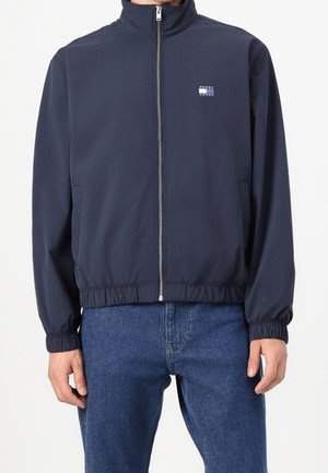 Granatowa kurtka na zamek z elastycznymi mankietami i ściągaczem u dołu, małe logo Tommy Jeans na lewej piersi, noszona z niebieskimi jeansami.