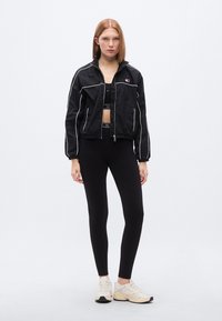 Μαύρο κοντό σακάκι με λευκές ρίγες, ελαστικά μανίκια και φερμουάρ κλεισίματος, συνδυασμένο με μαύρα κολάν και ανοιχτόχρωμα αθλητικά παπούτσια.