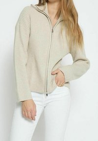 Beige ribbat zip-up tröja med huva, med långa ärmar och en avslappnad passform, bärs över vita byxor. Slät textur och minimala accenter.