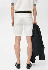 Weiße Denim-Shorts mit umgeschlagenen Säumen, zwei Gesäßtaschen und einem schwarzen Gürtel. Eine schwarze Jacke in der Hand, Sandalen an den Füßen.