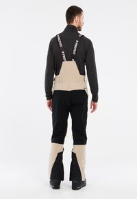Beige en zwarte sneeuwbroeken met verstelbare banden, ritszijden en een gestructureerde afwerking. Ontworpen voor winterweer en actieve beweging.